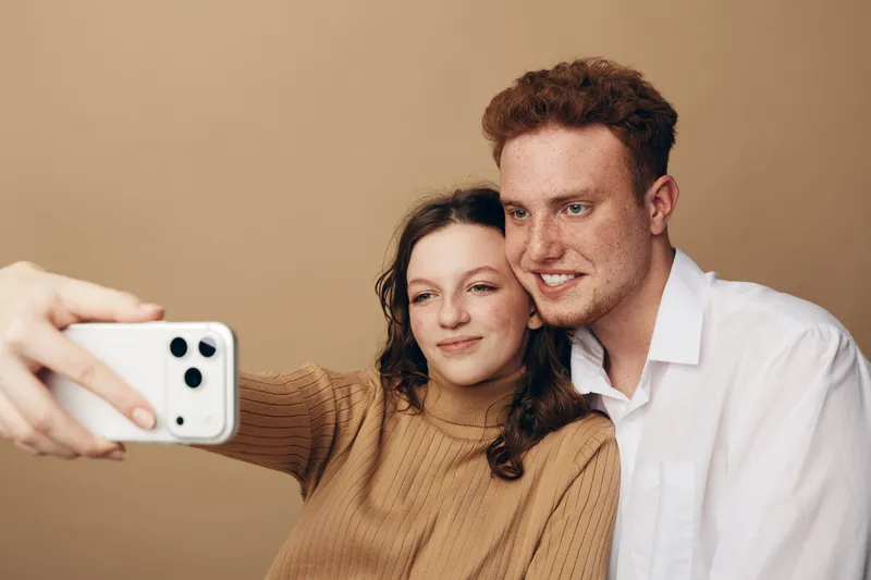 Young couple with curly hair takes a selfie together on smartphone. They smile and pose closely in casual clothes against a plain beige background. Concept of friendship and modern technology.