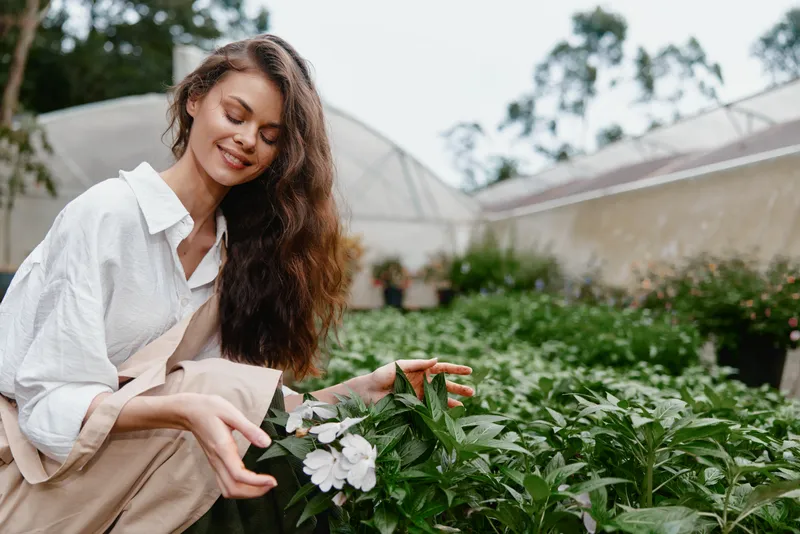 Young woman enjoying nature while gardening with flowers, wearing casual clothing, surrounded by greenery in a serene greenhouse setting