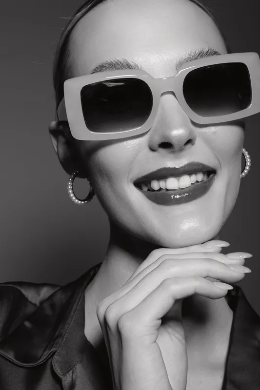 Young white woman with pale skin wearing oversized sunglasses and pearl earrings smiles confidently. Black and white close-up portrait with stylish mood and modern fashion concept.