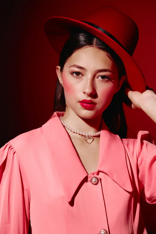 Woman with flawless bright makeup wearing an oversized coral jacket and maroon hat toned soft studio light creates a fashion beauty creative mood concept.