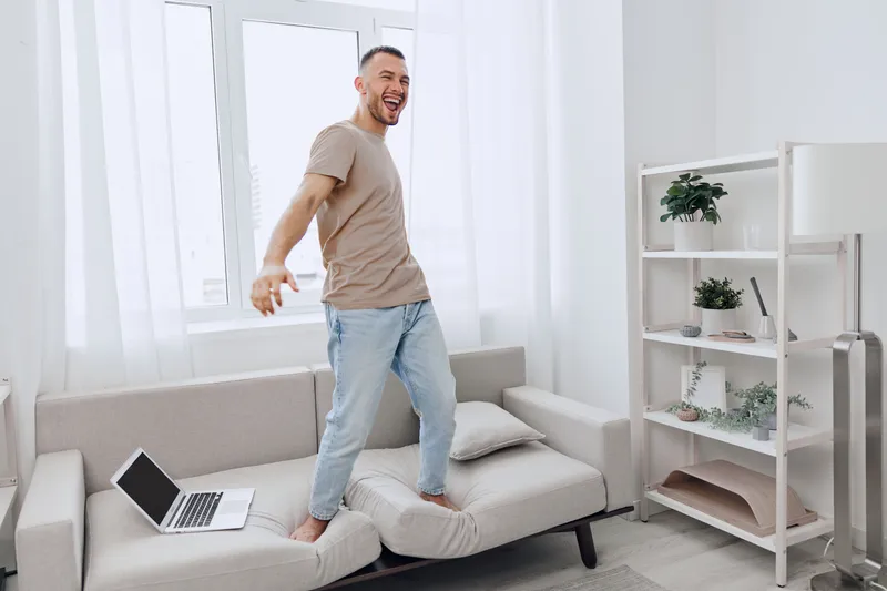 Energetic man expressing joy while standing on a couch, representing the blend of personal and professional life challenges in modern living today.