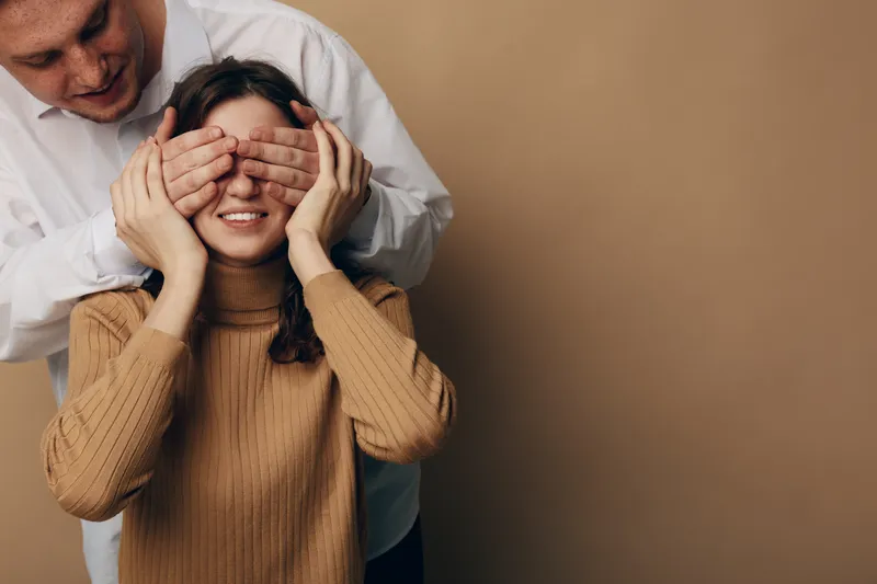 Young man covers eyes of smiling woman in brown sweater, playful moment between couple on beige background. Concept of surprise, love, relationship, and happiness in casual setting.