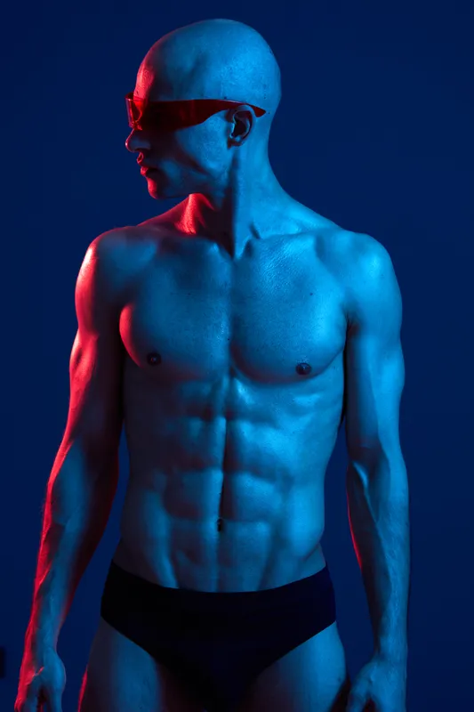Athletic man posing in artistic lighting, showcasing his fit physique and intense expression, captured in vibrant blue and red tones for a dynamic effect.