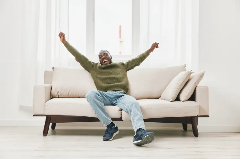 Cheerful African man celebrating with raised arms on a light couch in a bright airy room, dressed casually in a green sweater and jeans for a relaxed atmosphere