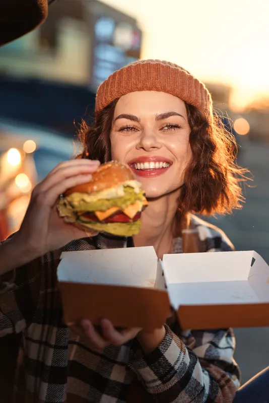 Woman holding a burger outdoors, smile and candid takeout lifestyle portrait in goldenhour with cozy beanie and plaid, authenticity, emotional storytelling and mindful living capture the moment.