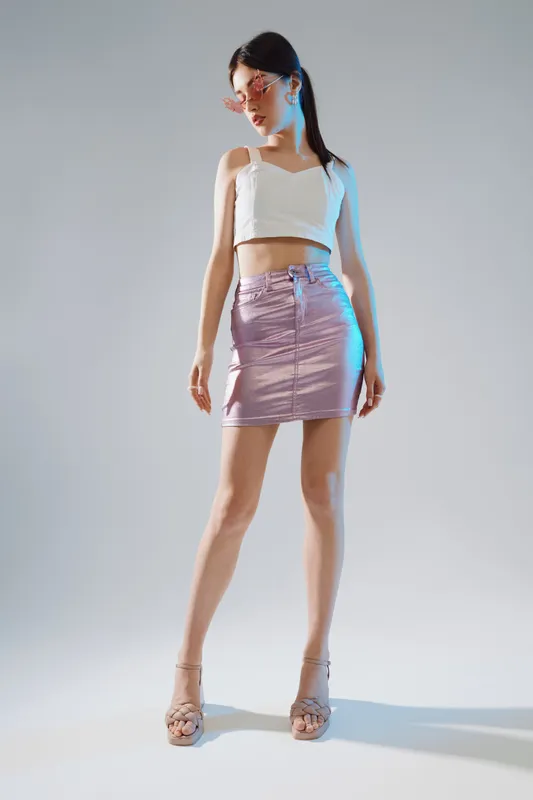 Young woman in fashionable outfit poses confidently wearing a white crop top and metallic pink skirt. Studio shot with soft lighting highlights beauty and fashion concept.