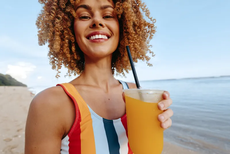 A joyful young woman with curly hair drinks orange juice on a sunny beach, smiling as the sea glitters in the background, capturing a carefree moment of relaxation while a colorful striped swimsuit