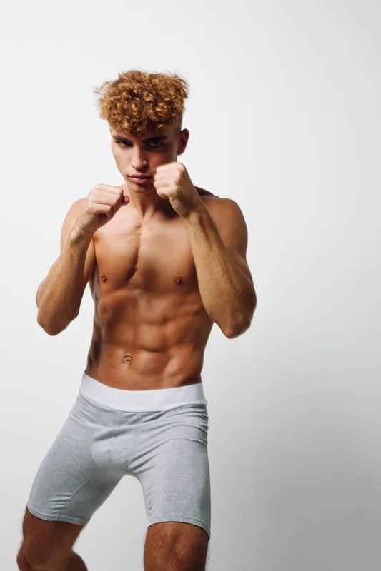 Athlete, male and fight in preparing boxing stance with hands raised and strong confident expression on bodybuilder in shorts against white background. Fit sportsman readies for workout