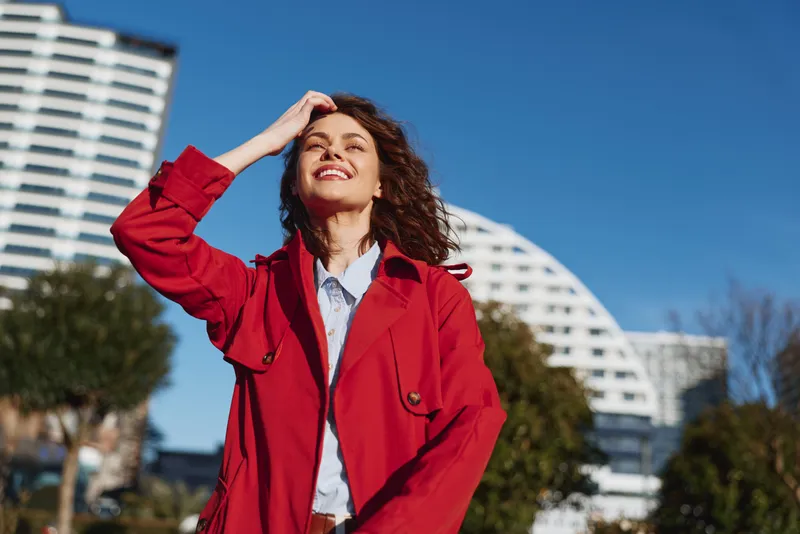 One person, young woman, outdoors on a sunny day wearing a red jacket smiles confidently against modern city buildings, radiating optimism and energy throughout day.