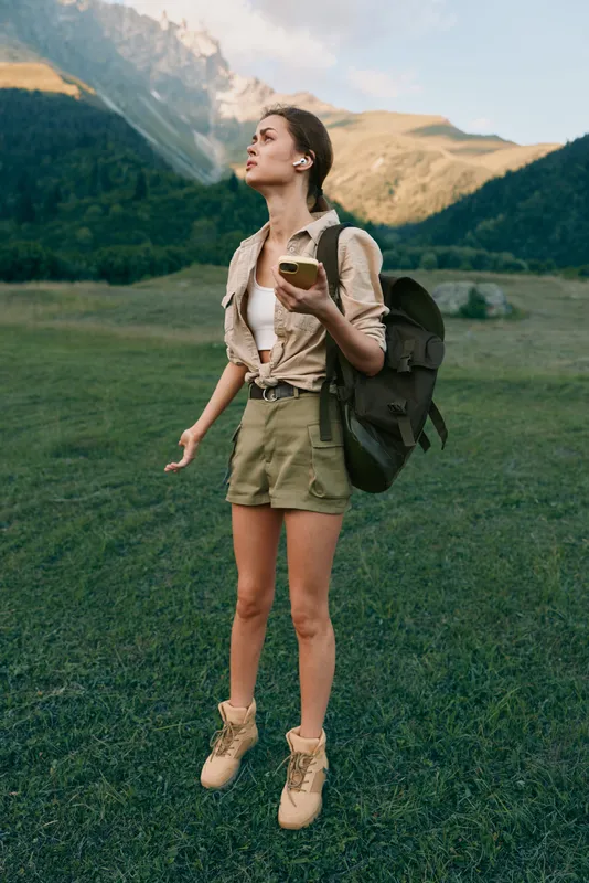 Young woman with backpack hiking in mountains holding an apple, wearing shorts and boots. Outdoor lifestyle, adventure, nature, healthy living, travel concept.