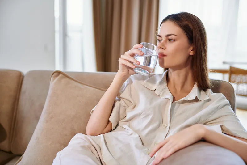Woman drinking water from a glass on a sofa at home, casual portrait showing relaxation, hydration and comfort in living room, pajama lifestyle and peaceful domestic moment