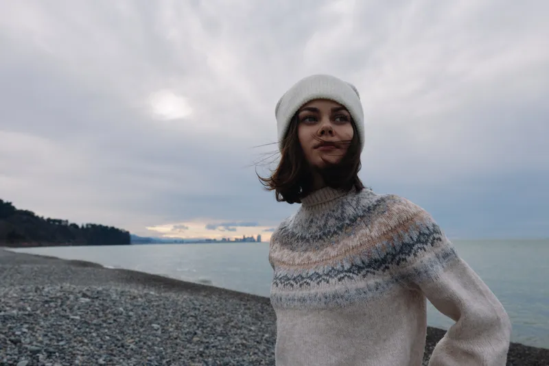 Woman on a pebble beach wearing a cozy sweater and wool hat, by the sea under a cloudy sky with a calm horizon and winter mood.