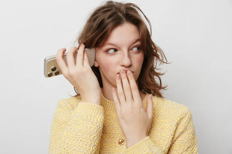 Young woman in yellow sweater holds smartphone near ear and covers mouth with hand, looking surprised. Concept of secret conversation, gossip, and curiosity in casual lifestyle.