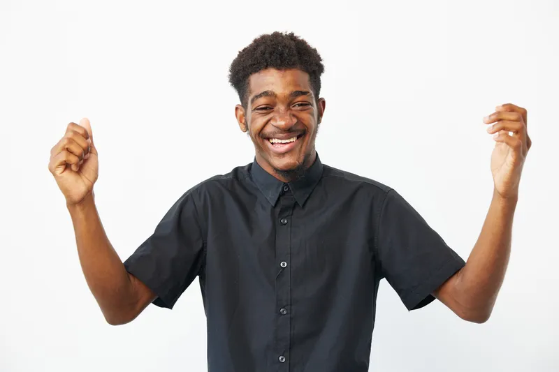 Happy black man celebrating with raised hands, wearing a stylish black shirt. His joyful expression radiates positivity and enthusiasm, perfect for uplifting themes.