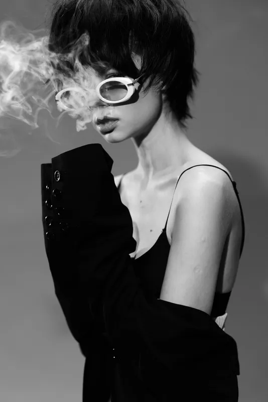Black and white portrait of a stylish woman with short hair wearing sunglasses and smoking, exuding a moody and edgy vibe.