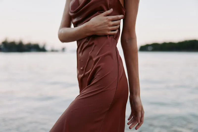 A close shot of a person in a brown dress standing by a calm lake, hands resting on the waist, soft light, gentle breeze, reflective water, serene mood.