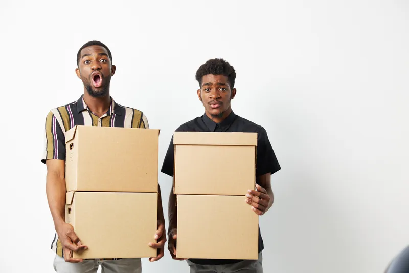 Two surprised African American men holding cardboard boxes, showcasing expressions of excitement and curiosity. Ideal for concepts of storage, delivery, or surprise.