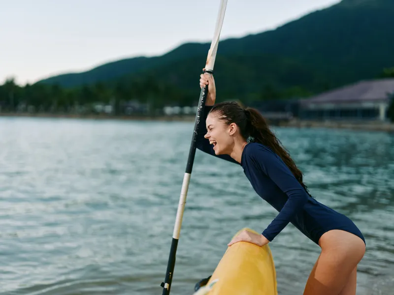 Smiling woman in navy swimsuit enjoys kayaking on calm water near tropical island with mountains in the background during daytime adventure.