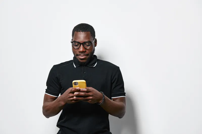African American man with dark skin wearing glasses and black polo shirt using smartphone for texting or browsing on white background. Casual, technology and communication concept.