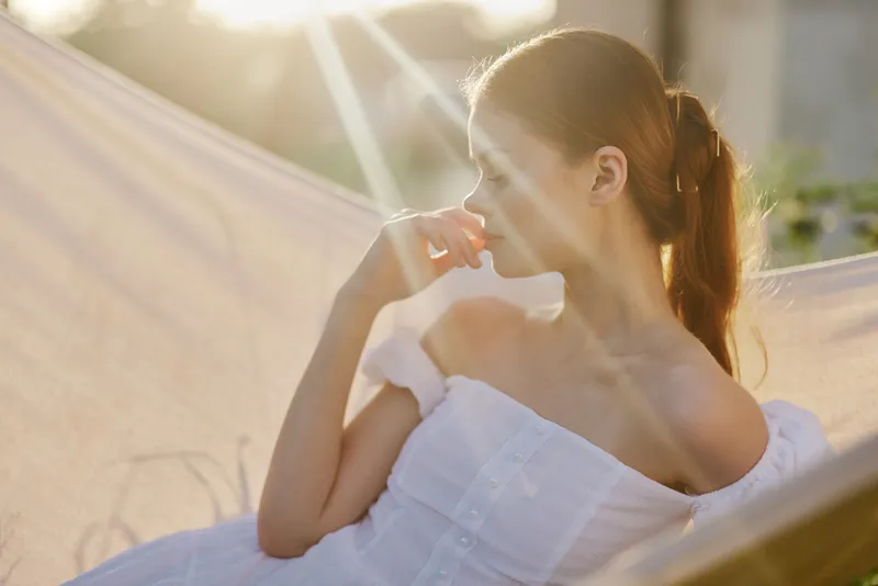 Young woman enjoying a sunny afternoon in a white dress, peaceful expression, sun rays creating a warm glow, serene outdoor setting