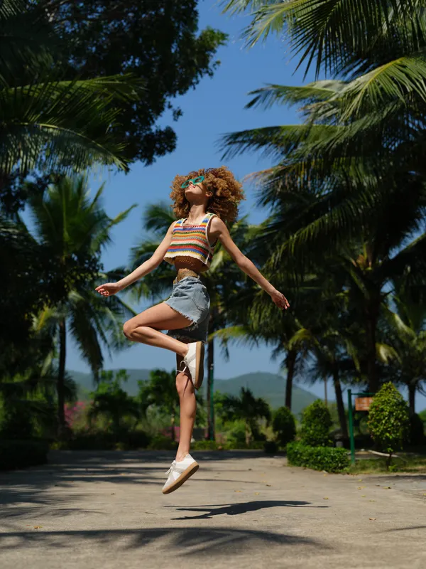joyful young woman with curly hair jumping outdoors on sunny day, wearing colorful striped top and denim shorts, surrounded by palm trees and clear blue sky