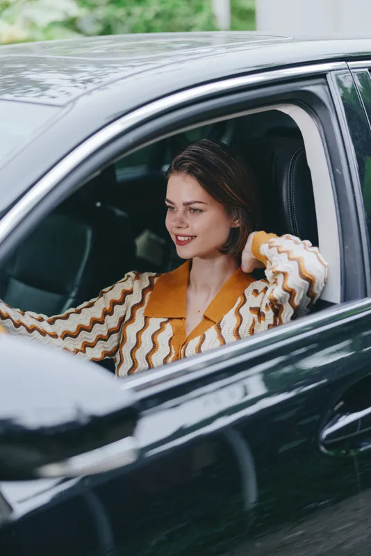 Woman sits in car, smiling, wearing a striped orange and white sweater, captured in a candid moment behind the wheel.