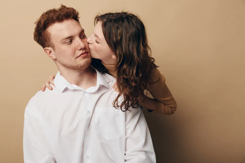 Young couple in casual clothes share intimate moment, woman kissing man on cheek. Beige background, warm lighting, concept of love, affection, and close relationship.