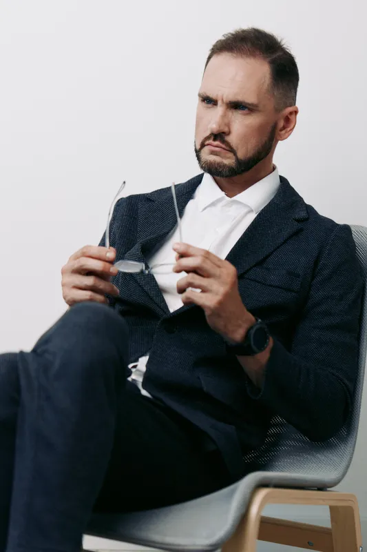 Professional business man sits with a thoughtful, focused expression, adjusting his glasses on an isolated colored background, conveying calm confidence and leadership readiness.