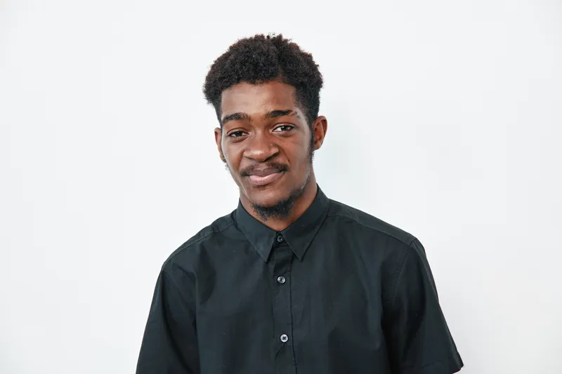 Young black man with dark skin wearing black shirt standing against white background looking at camera with slight smile and relaxed posture in studio portrait.