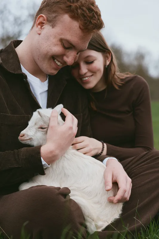 Loving couple outdoors with a gentle goat kid, sitting on grass and sharing smiles in a sunny field, showcasing affection, tenderness, and a warm countryside lifestyle with animals