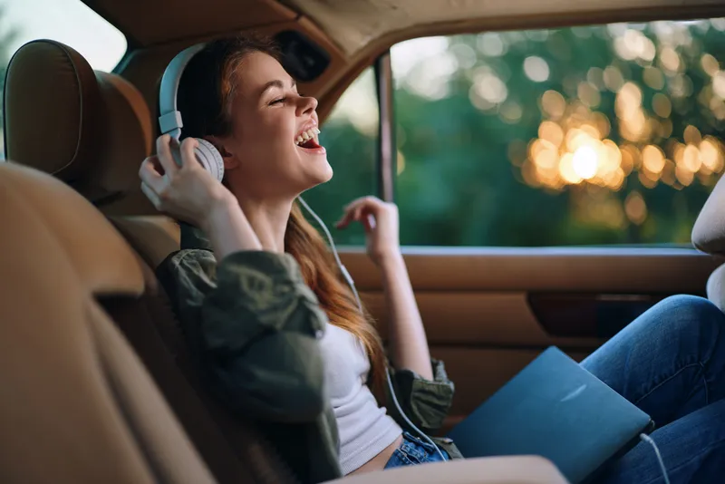 Happy young woman enjoying music with headphones in a car during sunset, wearing casual clothes and smiling joyfully.
