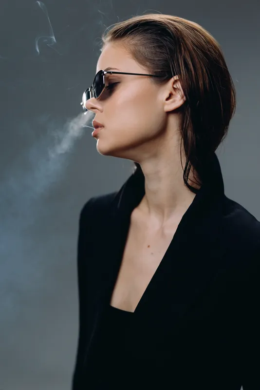 Stylish woman in sunglasses smoking cigarette with smoke on dark background, elegant black outfit, modern fashion portrait