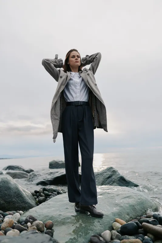 Young woman stands on rocky shore wearing wide pants and coat, looking confident and relaxed with hands behind head. Cloudy sky and sea background create calm natural atmosphere.