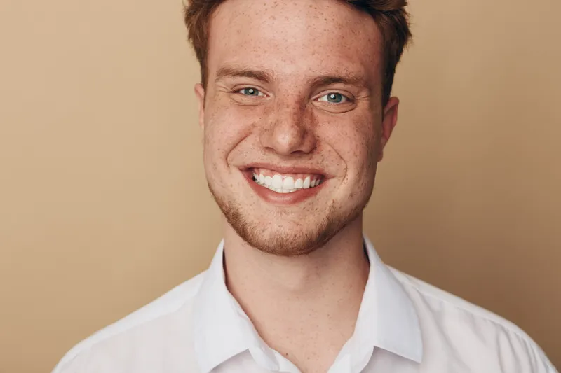 Smiling young man with red hair and freckles wears white shirt against beige background. Close-up portrait with natural light shows happy confident expression and healthy skin texture.