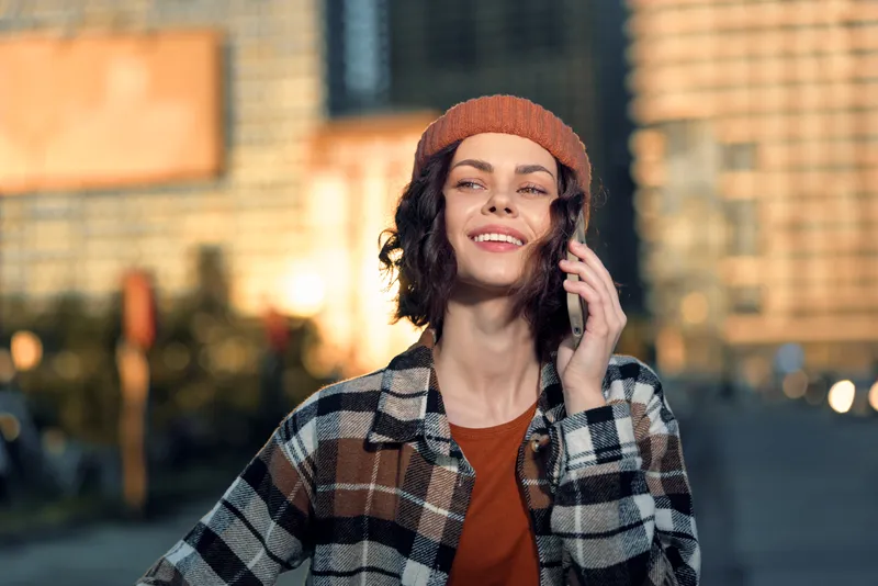 Woman smile phone urban candid authenticity golden glow lifestyle mindful living emotional storytelling portrait of a young adult in beanie and plaid jacket enjoying city sunlight.