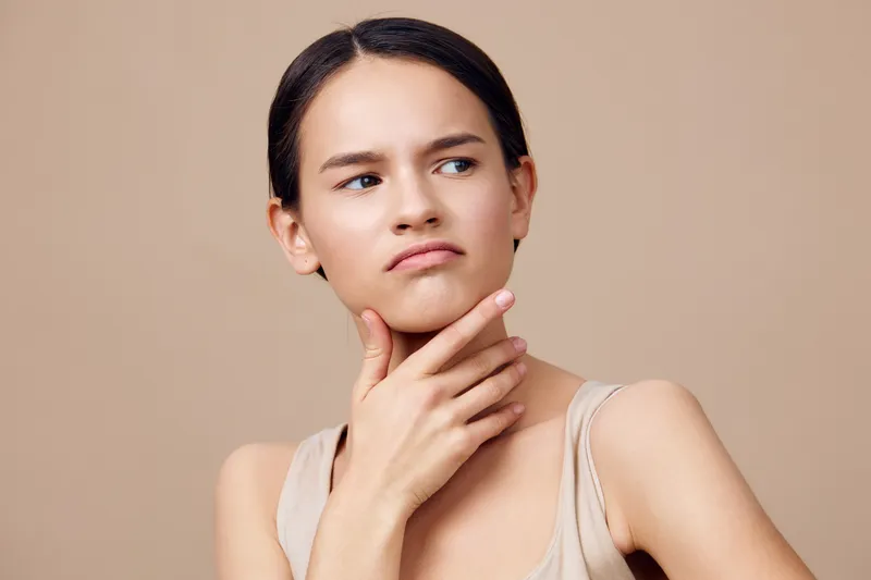 Thoughtful young woman with an expressive face contemplating options against a plain beige background, reflecting confusion and intrigue in a minimalistic style