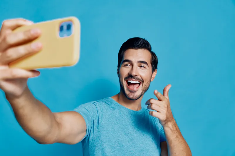 Cheerful smiling man taking selfie with smartphone against a bright blue background. Happy lifestyle and technology concept for social media enthusiasts.