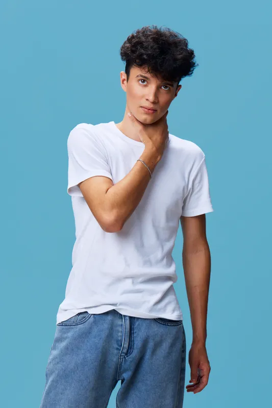 Young man with curly hair wearing white t-shirt and jeans stands against blue background looking thoughtful and calm, casual style, studio portrait.