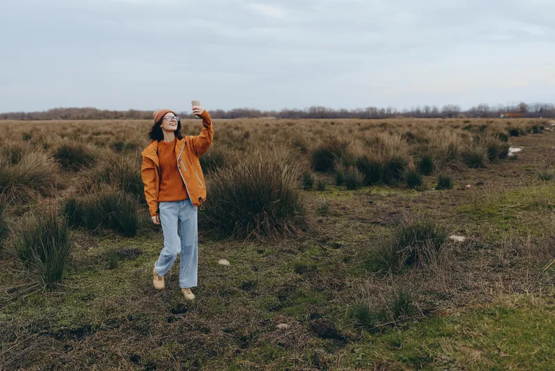 Woman outdoors walking field, wearing orange jacket and jeans, sunglasses, relaxed lifestyle, cloudy sky, natural grasses, rural landscape, candid moment, soft breeze, calm