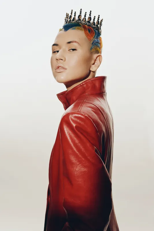 Young woman with colorful braided hair and crown wearing a red leather jacket, standing confidently against a plain white background in bold fashion style.