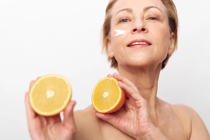 Mature woman with short hair applies facial cream and holds two halves of fresh orange in hands. Concept of natural skincare, healthy lifestyle, vitamin c, and beauty care for aging skin.