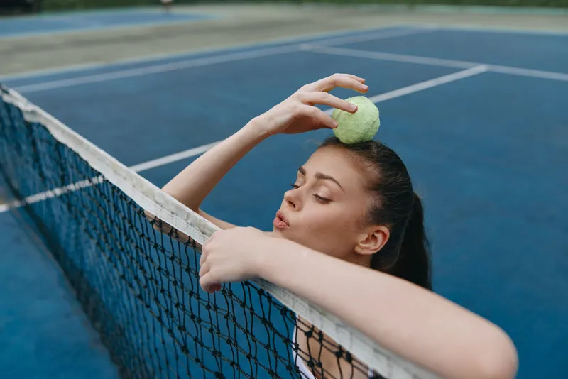 Young female tennis player in athletic wear balances a tennis ball on her head, showing focus and concentration on a blue court with a net, symbolizing sports dedication