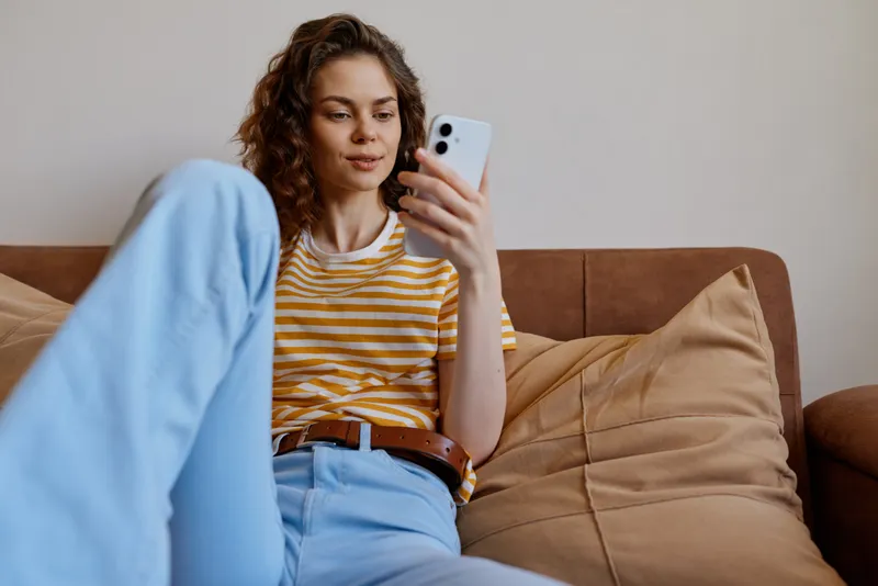Relaxed woman sits on a couch at home, looking at her smartphone, casual striped tee and light jeans. Cozy living room scene conveys casual lifestyle, comfort, and connection.