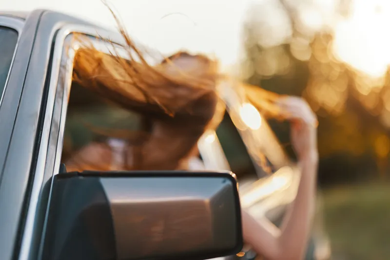 Young woman enjoying a carefree drive with hair flowing outside the window, golden hour sunlight illuminating the scene, vibrant emotions of freedom and joy.