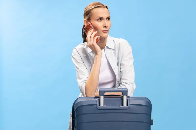 Young woman with suitcase talking on smartphone looks away thoughtful on blue background. Concept for travel, communication, planning, and business trip.