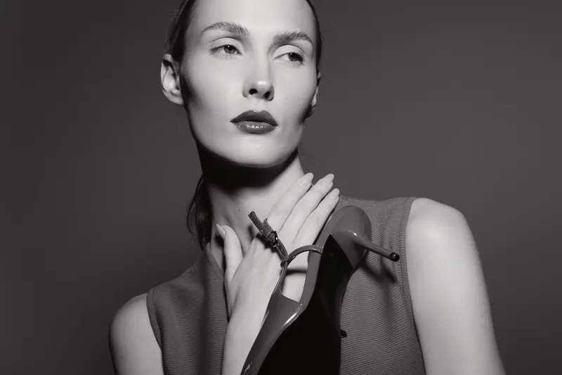 Elegant white woman with pale skin poses with high heel shoe on shoulder, looking away in black and white studio portrait.
