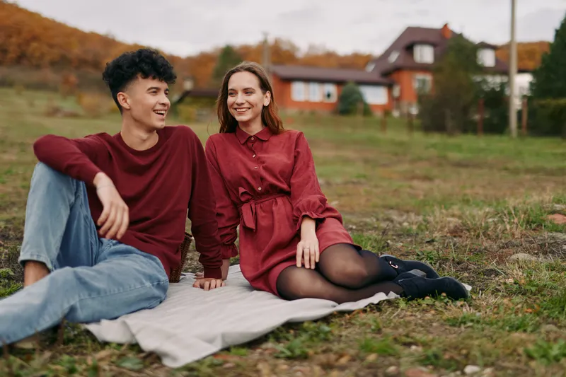 A genuine candid portrait of a young couple sitting on a blanket in a rural field, sharing warm smiles and natural interaction that conveys authenticity and reliable emotion.