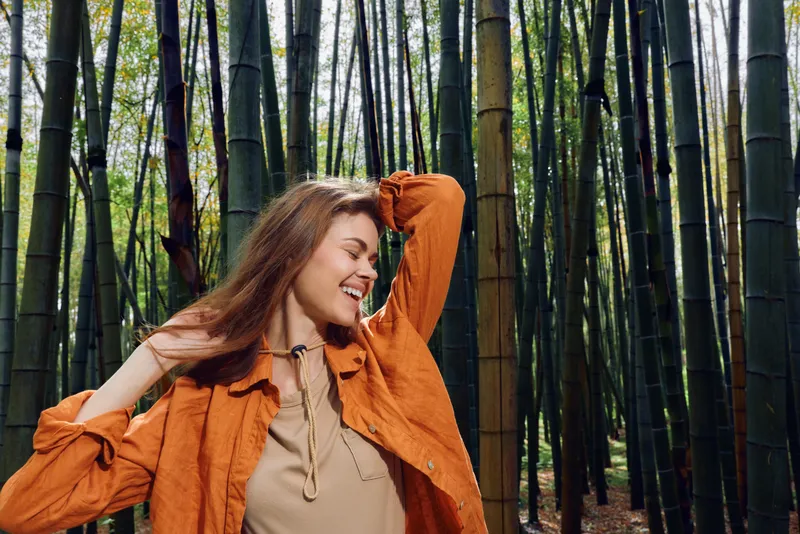 Woman in orange jacket smiling in bamboo forest, portrait of happy young woman enjoying nature and sunlight, relaxed pose and candid expression among tall green stalks