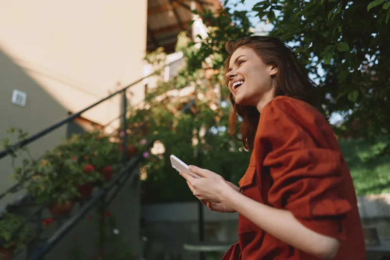 A smiling young woman in a red blouse enjoys a sunlit outdoor moment, holding a phone and laughing while leafy greenery frames a casual street setting, conveying candid lifestyle energy