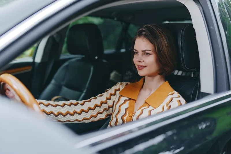 Portrait of a woman driving a car, wearing a striped orange blouse, seated in the driver's seat with a focused, calm expression.